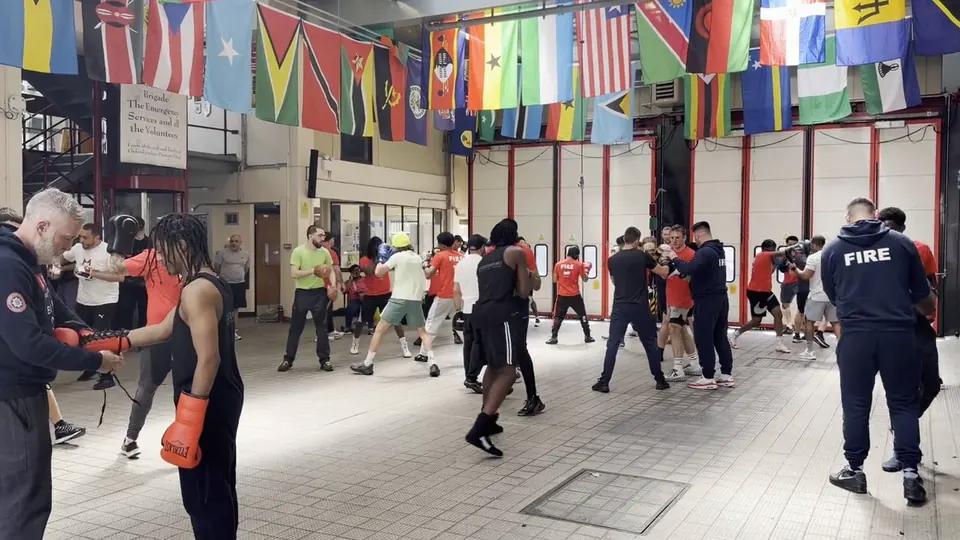 Video of a coach working the pads with a boxer as firefighters and boxers train in the fire station bay