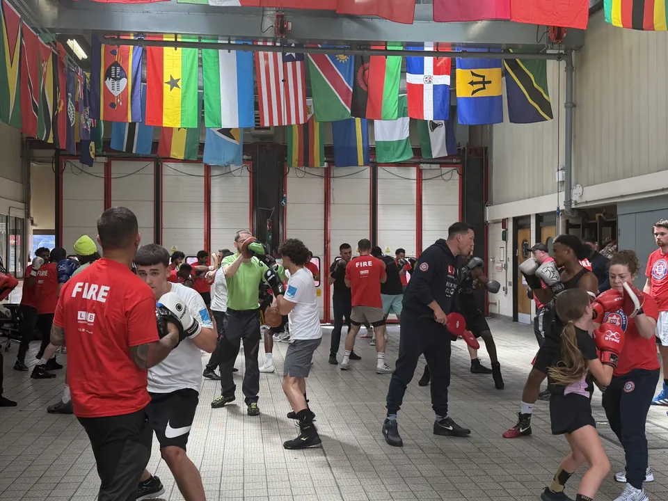 A firefighter in a red FIRE top holds pads for a boxer as coaches and young boxers train around them