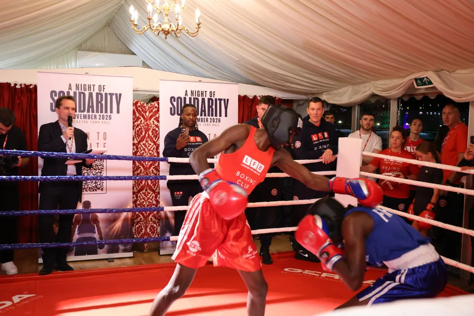 A boxer in red throws a punch as his opponent in blue ducks, with the ring announcer behind