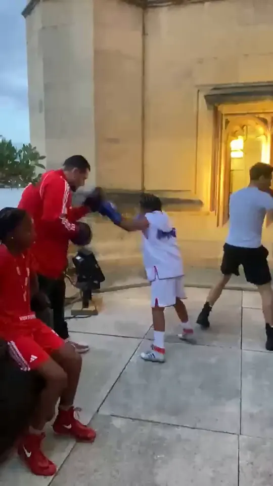 Video of a coach holding pads for a young boxer in a white vest in the evening light