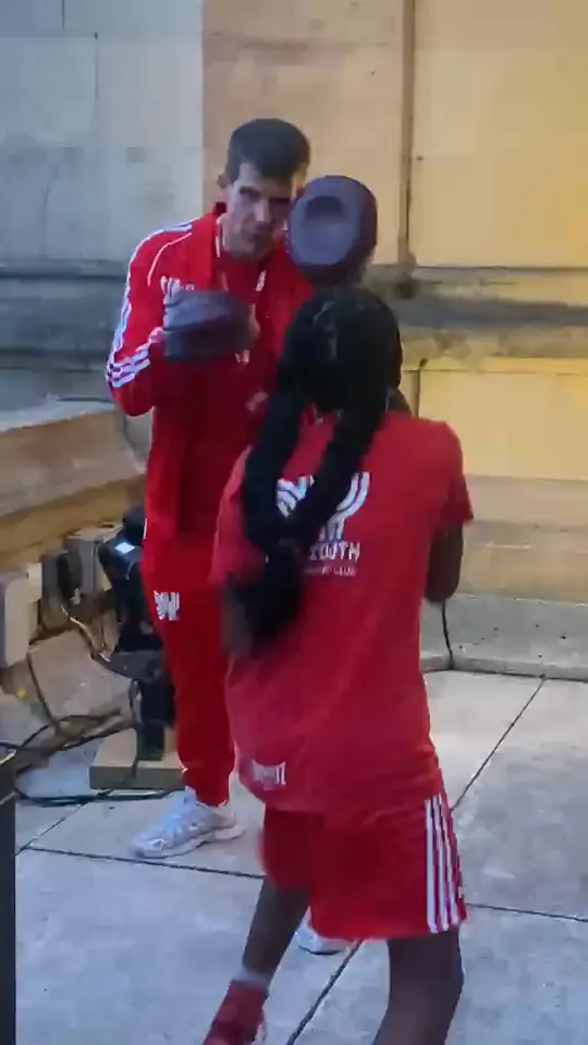 Video of a coach in a red tracksuit working pads with a young boxer on the terrace